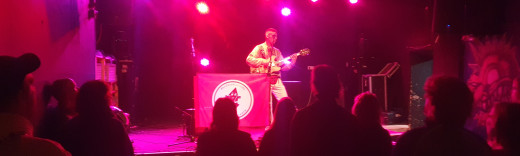 Vor einer Bühne in einem Konzertsaal mit einem Gitarristen stehen mehrere Personen, die ihm zuhören.