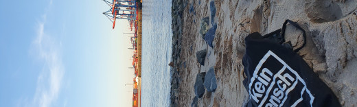 Am Elbstrand liegt ein Beutel mit dem Aufdruck "Kein Mensch ist illegal". Im Hintergrund die Elbe und der Hamburger Containerhafen.