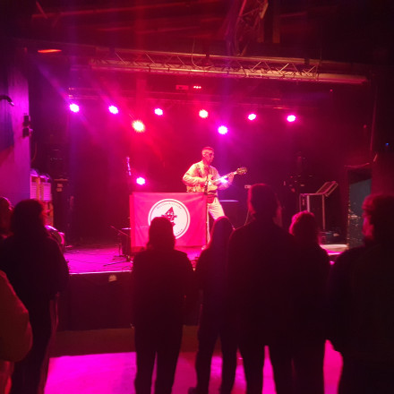 Vor einer Bühne in einem Konzertsaal mit einem Gitarristen stehen mehrere Personen, die ihm zuhören.