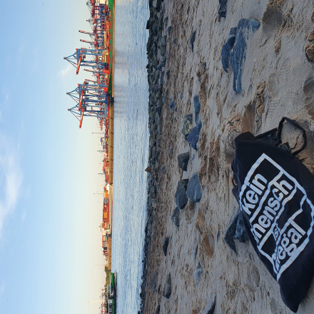 Am Elbstrand liegt ein Beutel mit dem Aufdruck "Kein Mensch ist illegal". Im Hintergrund die Elbe und der Hamburger Containerhafen.